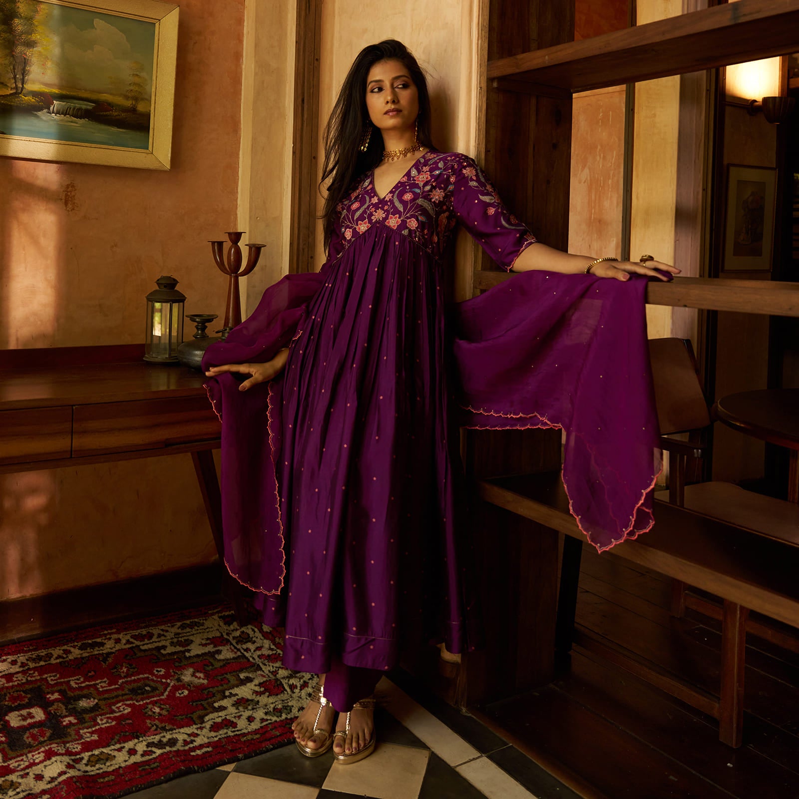 A woman models a traditional Indian outfit consisting of a deep purple long kurta with floral embroidery and a matching sheer purple dupatta, posed in a warm, rustic interior setting.