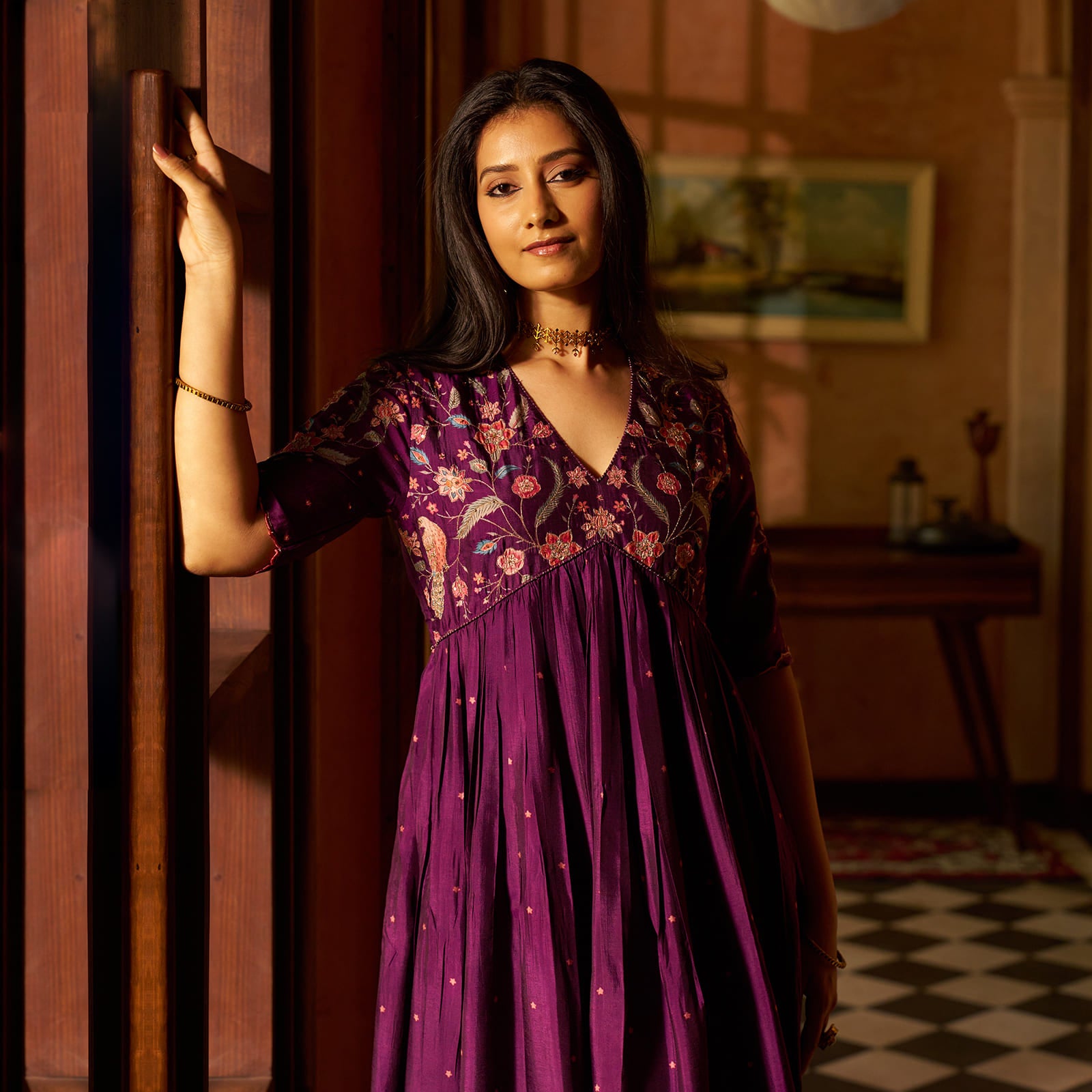A woman wearing a purple floral embroidered dress leans against a wooden door in a warmly lit indoor setting.
