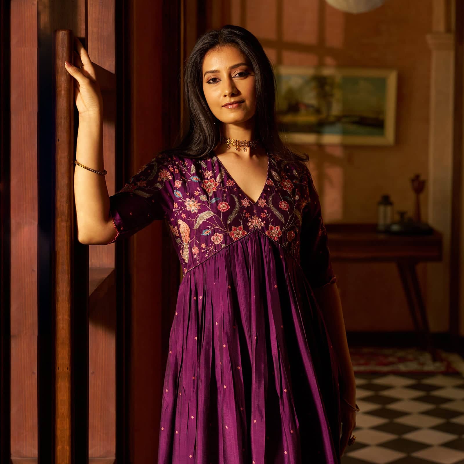 A woman wearing a purple floral embroidered dress leans against a wooden door in a warmly lit indoor setting.