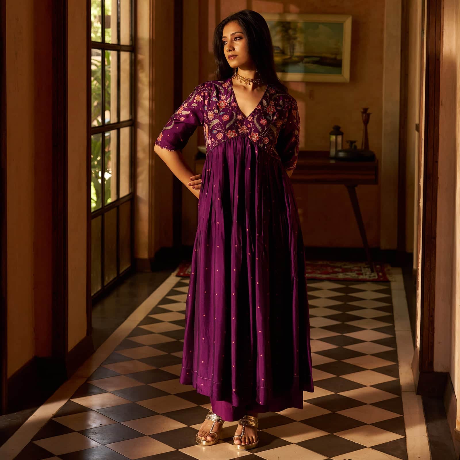 A woman models a purple traditional Indian dress with floral embroidery on the bodice and scattered gold zari embellishments on the skirt, standing in a hallway with a checkered floor. | 